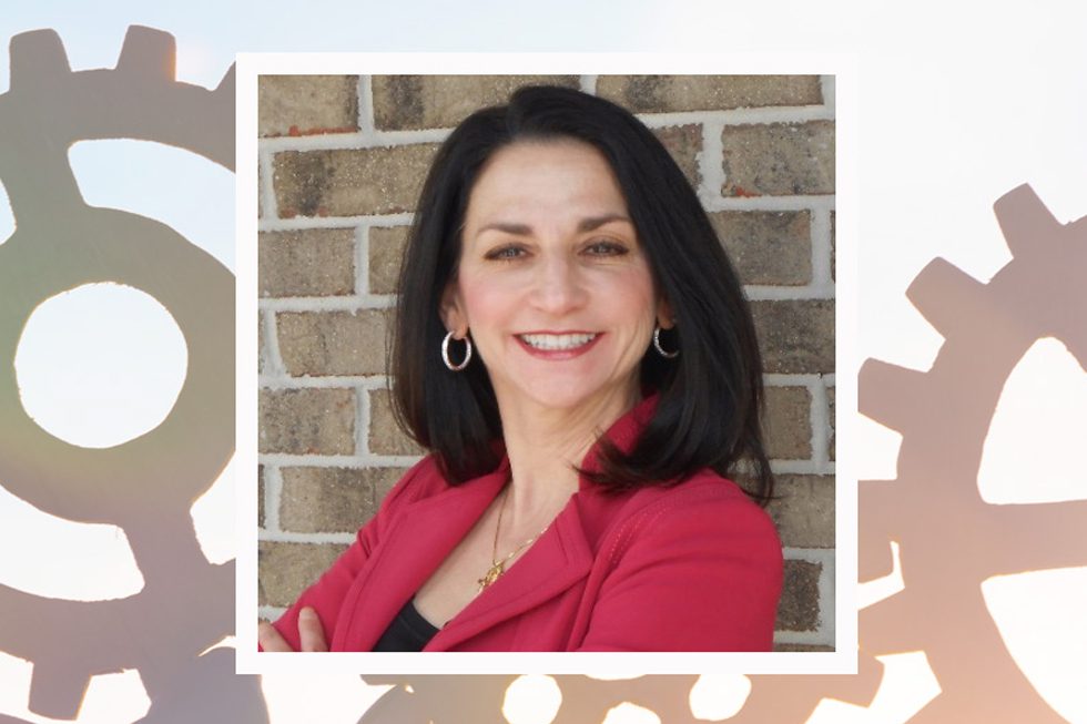 Smiling woman with shoulder-length dark hair in a pink blazer stands against a brick wall.